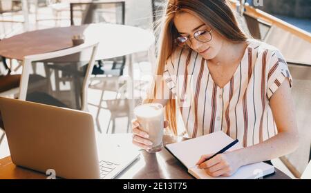 Chiudi foto di una donna caucasica con frettole e rosso capelli che indossano gli occhiali e che bevono una scrittura del cocktail in un prenotare e utilizzare un computer portatile nel Foto Stock