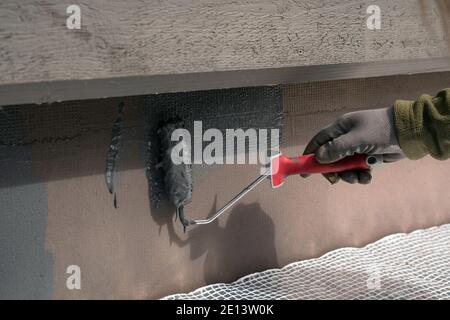 Uomo che dipinge casa socle con rullo di vernice. Primo piano della vernice grigia arrotolabile sulla base dell'edificio. Ristrutturazione domestica Foto Stock