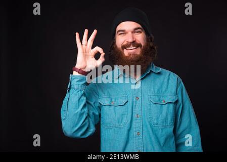 Una bella foto di un giovane uomo felice sopportato sorridendo alla telecamera e mostrando il segno ok vicino ad una parete scura . Foto Stock