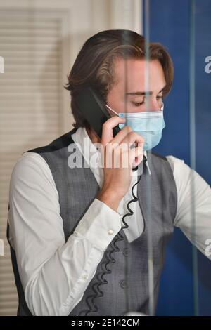 un giovane receptionist maschile attento che indossa una maschera chirurgica mentre si parla al telefono Foto Stock