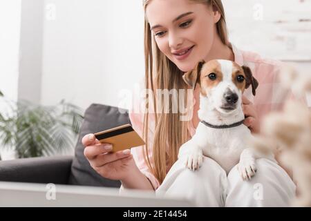 felice giovane donna che tiene la carta di credito vicino jack russell terrier in soggiorno Foto Stock