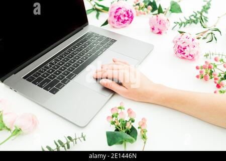 Donna che lavora su un computer portatile. Spazio di lavoro con mani femminili, laptop e fiori rosa su sfondo bianco. Lavoro remoto a casa Foto Stock