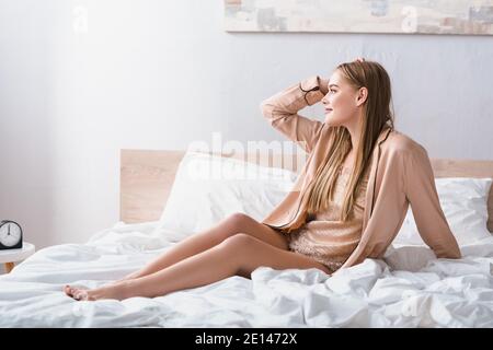 gioiosa giovane donna che guarda via mentre si siede a letto dentro camera da letto moderna Foto Stock