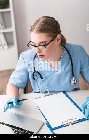 giovane veterinario in guanti di lattice utilizzando il computer portatile vicino a clipboard su tabella Foto Stock