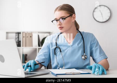 giovane veterinario in occhiali e guanti in lattice utilizzando laptop vicino appunti sul tavolo Foto Stock