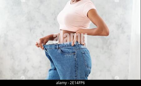 Donna nera irriconoscibile in jeans oversize che mostra i risultati della sua dieta di perdita di peso o liposuzione, sfondo leggero Foto Stock