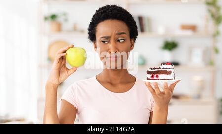 Giovane donna nera che guarda la mela con sospetto, non vuole mangiare frutta invece di torta a casa Foto Stock