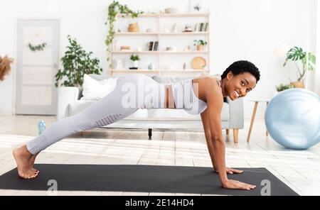 Bella giovane donna nera che fa l'addestramento domestico sul tappeto di yoga, in piedi in tavola, rinforzando i muscoli dell'abs a casa Foto Stock