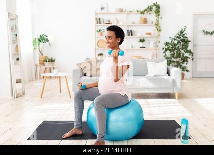 Donna nera incinta seduta sulla sfera di idoneità, che lavora fuori con i dumbbells nella sua palestra domestica Foto Stock