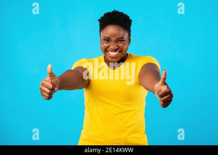 Plus-Size Black Woman gesturing Thumbs-Up con entrambe le mani, sfondo blu Foto Stock