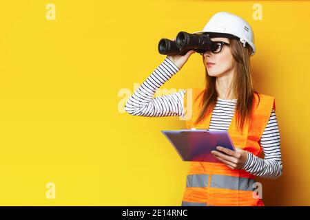 Giovane donna in gilet di segnale e casco da costruzione guardando attraverso binocoli su sfondo giallo. Foto Stock