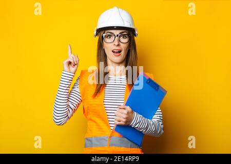 Giovane donna con un volto sorpreso in un gilet e elmetto tiene una clipboard su uno sfondo giallo. Concetto di costruzione, nuovo edificio. Banner. Foto Stock