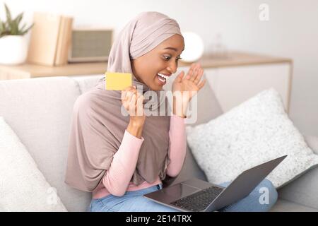 Donna nera eccitata in hijab con la carta di credito felice su grande vendita in negozio online, shopping sul computer portatile a casa Foto Stock