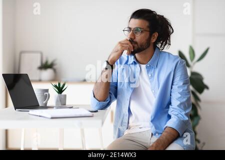 Premuroso giovane uomo arabo freelance seduto alla scrivania a casa, guardando da parte Foto Stock