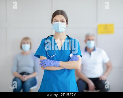 Ritratto di giovane dottore in uniforme e maschera facciale con pazienti in attesa di vaccinazione con coronavirus in clinica Foto Stock