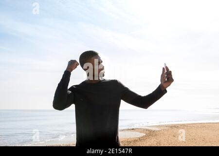 Giovane sportivo nero sulla spiaggia che mostra la sua forza mentre fa una videochiamata con il suo cellulare. Foto Stock