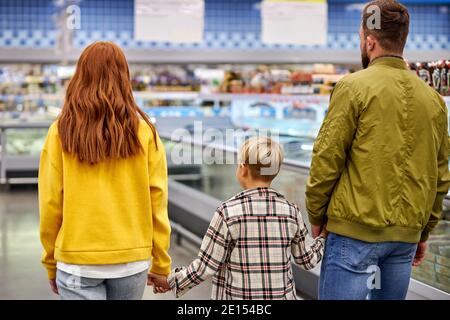 la famiglia cammina nel negozio di alimentari, si passa davanti a vetrine e scaffali con cibo, vista posteriore, in abbigliamento casual, andare avanti e acquistare Foto Stock