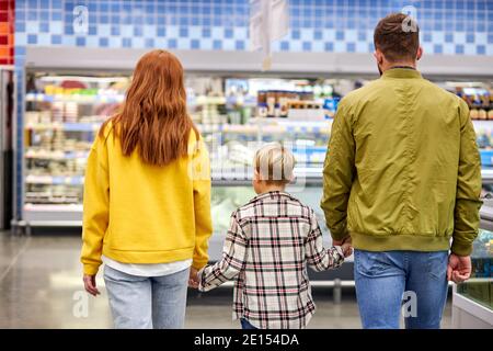 famiglia con il figlio che cammina nel negozio di alimentari, tenendo le mani insieme, si oltrepassano vetrine e scaffali con cibo, vista posteriore, in abbigliamento casual, andare fo Foto Stock