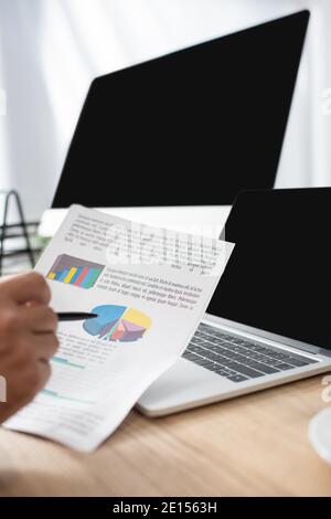 vista ritagliata del trader che punta con la penna alle infografiche vicino al laptop e monitor con schermo vuoto, primo piano sfocato Foto Stock