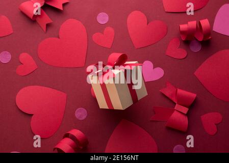 Carta arte banner concetto di San Valentino con confezione regalo fatta a mano, nastro tagliato di carta, arco, e un sacco di cuori su sfondo rosso con spazio per il testo. Foto Stock
