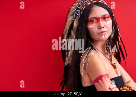 donna sciamanica indiana con sciamano guerriero si compongono isolati in studio su sfondo rosso, dipinti colorati sul viso e il corpo, lei sta guardando ca Foto Stock