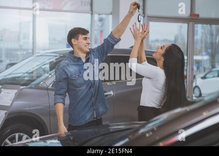 Coppia felice che si diverte insieme al salone della concessionaria, dopo l'acquisto di una nuova automobile insieme. Allegro bell'uomo che ride tenendo in mano le chiavi dell'auto, Ciao Foto Stock