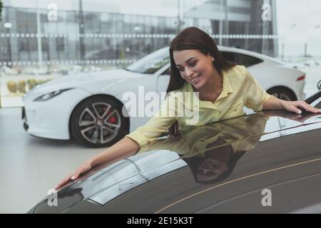 Giovane donna emozionata sorridente, abbracciando la nuova auto in vendita presso la concessionaria. Bella donna che acquista la sua prima auto al salone di veicoli. Allegro femmina driv Foto Stock