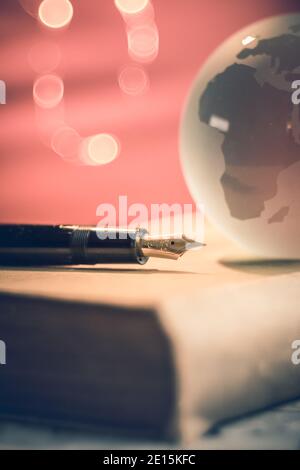 primo piano della penna nera e del globo di vetro con i paesi su un vecchio libro. Sfondo arancione illuminato con piccole luci spot. Concetto di scrittura o viaggio Foto Stock