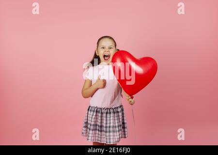 primo piano entusiasta rapturous estatica ragazza in abito rosa tiene rosso palloncino a forma di cuore e dà pollici in su isolato su sfondo rosa Foto Stock