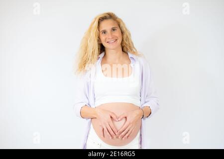 Donna incinta sorridente che fa un gesto del cuore con le dita su sfondo bianco. Concetto di gravidanza, maternità, preparazione e attesa. Foto Stock