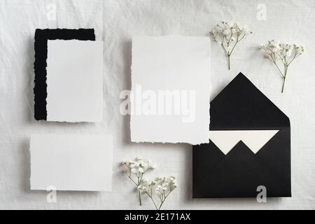 Mokup femminile di cancelleria per matrimoni su sfondo in tessuto bianco pastello. Fiori di Gypsophila, carte bianche, busta nera vista dall'alto. Foto Stock