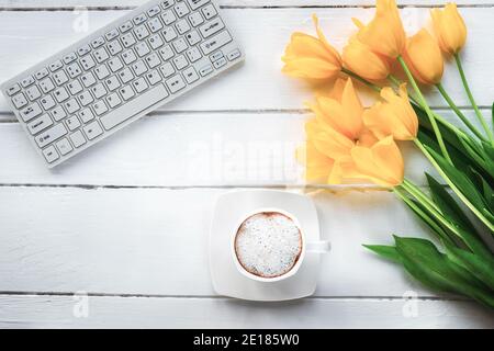 Tulipani gialli, tastiera e tazza di cappuccino su un tavolo di legno bianco. Concetto di Giornata della Donna. Vista dall'alto, disposizione piatta. Foto Stock