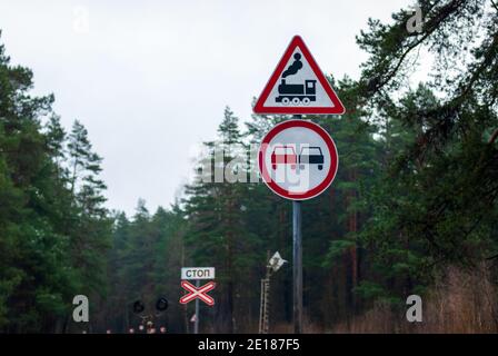 Segnaletica stradale per attraversamento ferroviario, nessun sorpasso e segnale di stop (fermata scritta in russo) contro alberi di foresta su strada di campagna Foto Stock