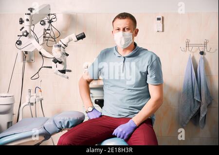 Ritratto del dentista uomo in guanti sterili e maschera medica guardando la macchina fotografica. Stomatologo maschile seduto vicino al microscopio dentale. Concetto di odontoiatria e stomatologia. Foto Stock