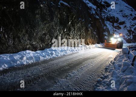 lo spazzaneve passa con le luci attraverso le strade di montagna innevate di sera per ripulire il traffico stradale Foto Stock