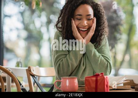 donna afro-americana stupita guardando rosso scatola regalo di natale vicino a cup in caffetteria Foto Stock