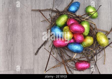 Uova di pasqua multicolore avvolte in foglio di colore rosa, verde, blu e giallastro in un nido naturale, di bastoni e ramoscelli, su uno sfondo grigio di legno bianco. Foto Stock