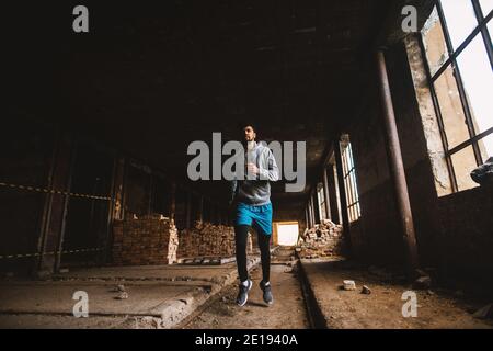 Ritratto di un giovane afro-americano attivo e motivato attraente atletico con auricolari che corrono all'interno del luogo abbandonato. Foto Stock