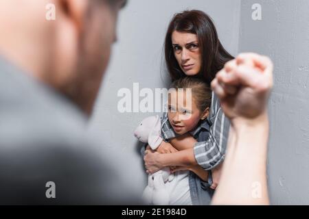 Donna spaventata abbracciando figlia con lividi e giocattolo morbido vicino muri e marito che mostrano il pugno su un primo piano sfocato Foto Stock