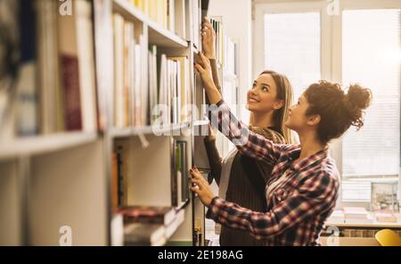 Due giovani ragazze studentesche sorridenti che scelgono i libri dalla libreria della scuola. Foto Stock