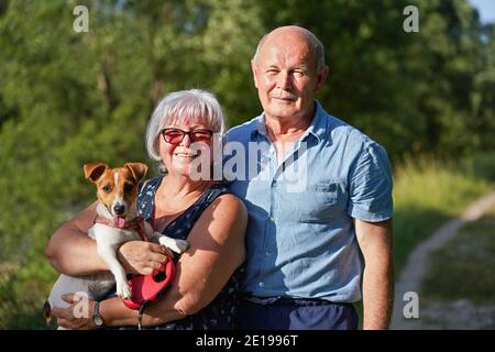 Coppia di anziani che posano con il loro cane Jack Russell piccolo, lingua fuori. La donna la tiene sulle mani, gli alberi offuscati e lo sfondo della strada di campagna Foto Stock