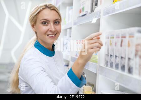 Splendida giovane e felice farmacista femminile in camice bianco che organizza i prodotti sugli scaffali, sorridendo alla macchina fotografica. Allegro chimico godeign lavorare a lui Foto Stock
