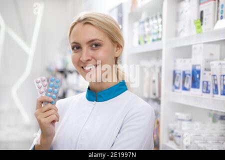 Primo piano di una bella e felice giovane farmacia sorridente alla macchina fotografica, tenendo blister di pillole. Sicuro farmacista femminile che vi offre vitamina p Foto Stock