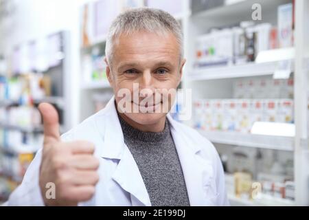 Allegro farmacista senior sorridendo gioiosamente, mostrando pollici in su, copia spazio. Felice chimico maschio maturo in camice bianco che mostra i pollici in su, lavorando a t Foto Stock