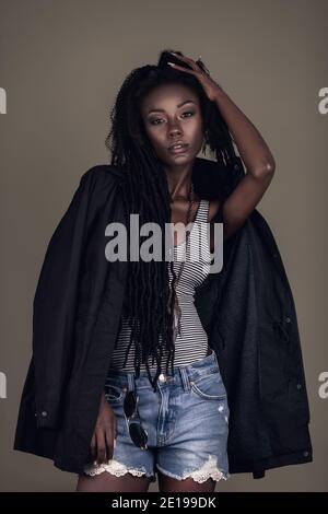 Ritratto di una giovane femmina nera con lunghi dreadlock, bel trucco e labbra morbide che si posano da sola indossando gioielli e pantaloncini in denim. Foto Stock