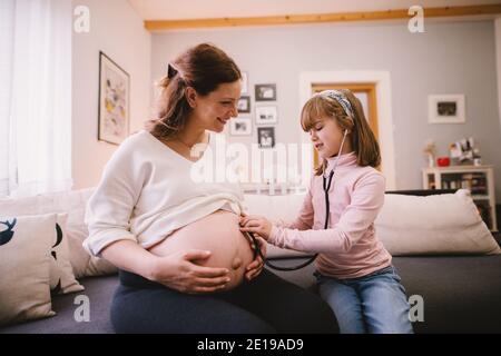 Piccola ragazza graziosa che ascolta il ventre della sua madre incinta attraverso uno stetoscopio. Concetto di famiglia e nuova vita. Foto Stock