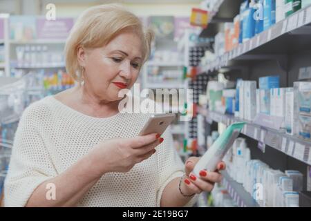 Donna anziana che usa il suo smartphone in farmacia, scannerizzando il codice qr di un prodotto. Anziana cliente di sesso femminile navigando online descrizione di un medico p Foto Stock