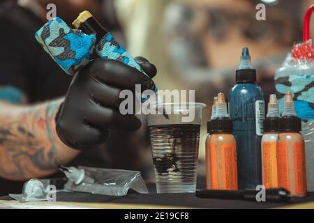 Primo piano corto di inchiostro tatuaggio che si diffonde in un bicchiere d'acqua, artista tatuaggio che pulisce la sua macchina tatuatrice in un bicchiere d'acqua, mentre lavora. Selettivo fo Foto Stock