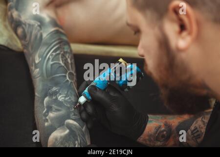 Un tatuista bearded che lavora nel suo studio, un braccio tattoing del suo cliente. Artista di tatuaggio professionale che utilizza tatuaggio m. Foto Stock