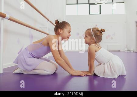 Due adorabili ballerine di ragazze che si siedono faccia a faccia, tenendo le mani, sorridendo l'un l'altro alla scuola di balletto. Bella ballerina giovane e la sua cute litt Foto Stock